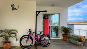 Ein pinkfarbenes Fahrrad steht auf einem Balkon mit einem Sandsack, Topfpflanzen und Blick zum Himmel.
