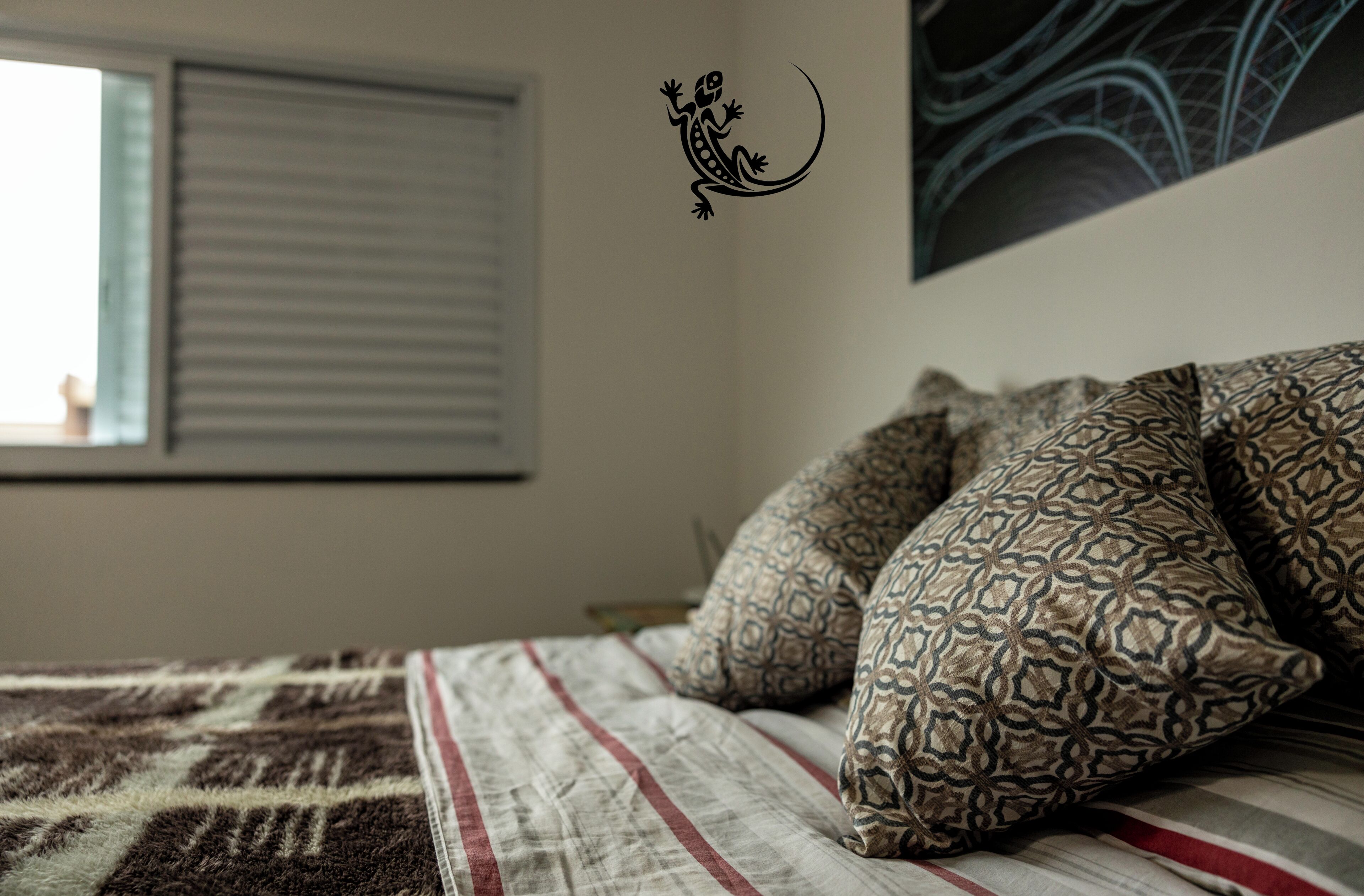 Ein Bett mit gemusterten Kissen und einer Streifen-Decke, ein Fenster mit Jalousien und eine Wandsticker-Echse.