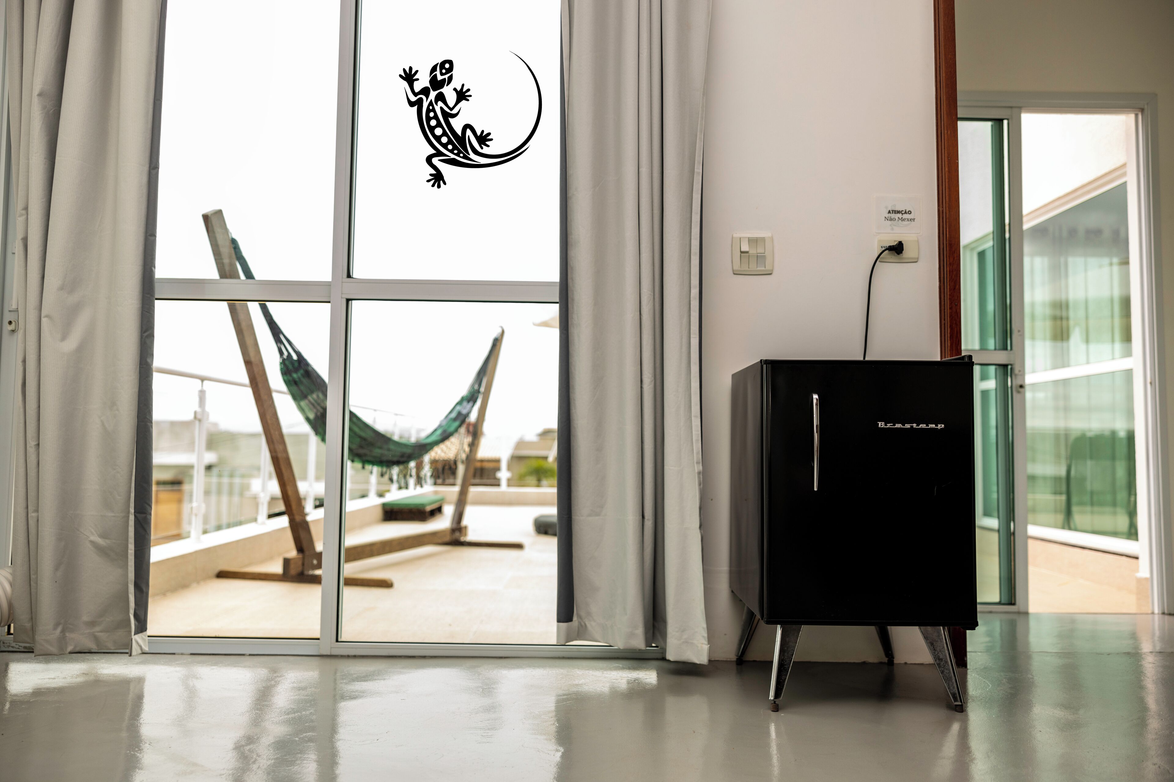 A room with a large window, a hammock, and a black refrigerator.