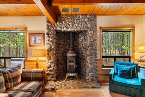 A cozy cabin interior with a stone fireplace, plaid furniture, and a view of the forest through large windows.