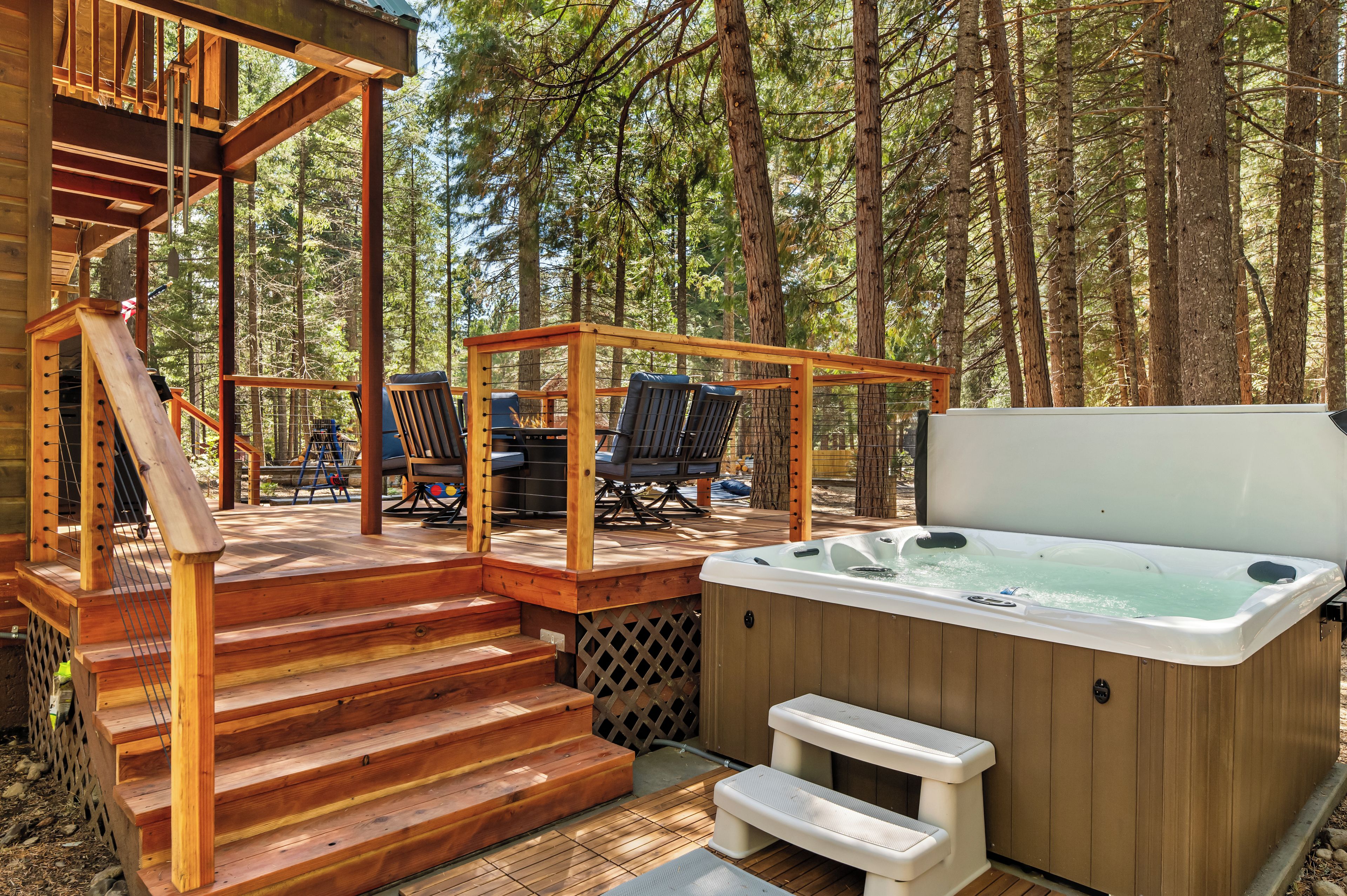 Een houten terras met een jacuzzi, buitenmeubels en een trap naar een overdekt gedeelte.