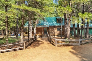 A rustic cabin with a green roof, surrounded by tall trees and a wooden fence.