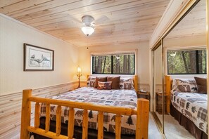 A wooden cabin bedroom with a bed, a framed picture on the wall, and a window with a forest view.