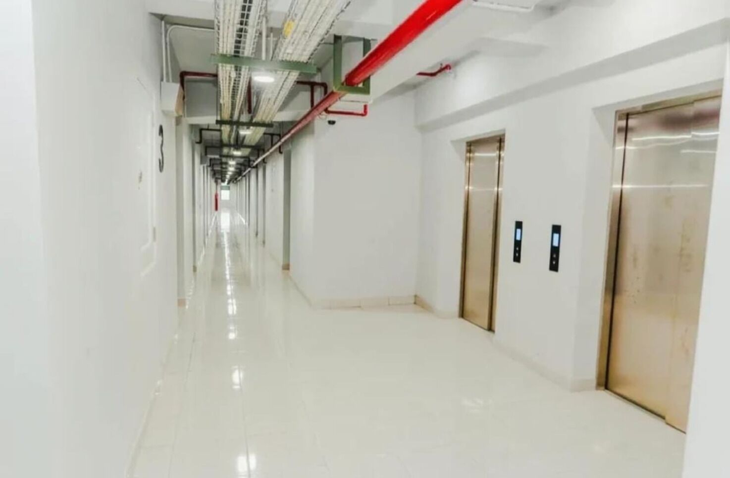 Un pasillo limpio y bien iluminado con piso brillante, dos ascensores y equipo visible de seguridad contra incendios.
