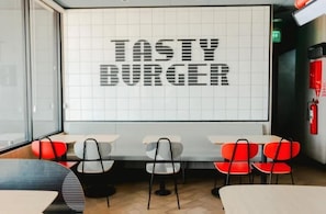 Et restaurantinteriør med et stort skilt, der siger 'TASTY BURGER' på væggen, borde med stole og en brandslukker.