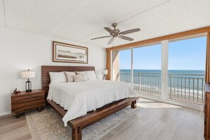Ein Schlafzimmer mit einem großen Bett, einem Nachttisch, einer Lampe, einem Deckenventilator, einem gerahmten Bild und Blick auf das Meer durch ein großes Fenster.