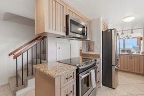 A kitchen with a microwave, oven, and refrigerator, wooden cabinets, granite countertops, and a view of the ocean through a window. - Oceanfront End-Unit Townhome with Two King Suites & Sweeping Coastal Views (New Smyrna Beach)