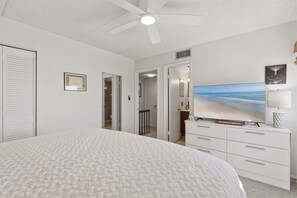 Quarto com cama, ventilador de teto, televisão e banheiro visível por uma porta aberta.