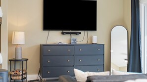 Ein Hotelzimmer mit Flachbildfernseher an der Wand, einem dunkelbraunen Schminktisch mit Schubladen, einem Nachttisch mit Lampe und einem großen Spiegel.