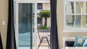 A view through a glass door into a patio area with a palm tree, outdoor furniture, and a building in the background. - Gulf Highlands Studio Getaway | King Bed, Bunks, Steps to the Beach (Panama City Beach)