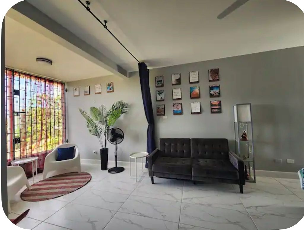 Un salon comprenant un canapé, des fauteuils, un ventilateur, une plante en pot et un mur orné de tableaux encadrés.