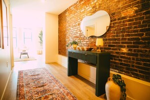 A hallway with a brick wall, a round mirror, a console table with a lamp, and a patterned rug. - Free Parking - Historic 2BR Apartment DT Halifax (Halifax)