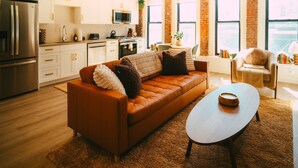Una sala de estar moderna con un sofá de cuero marrón, una mesa de centro de madera y una cocina con armarios blancos y electrodomésticos de acero inoxidable.