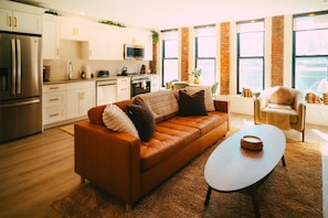 Un salon moderne avec un canapé en cuir marron, une table basse en bois et un coin cuisine doté de meubles blancs et d’appareils électroménagers en acier inoxydable.