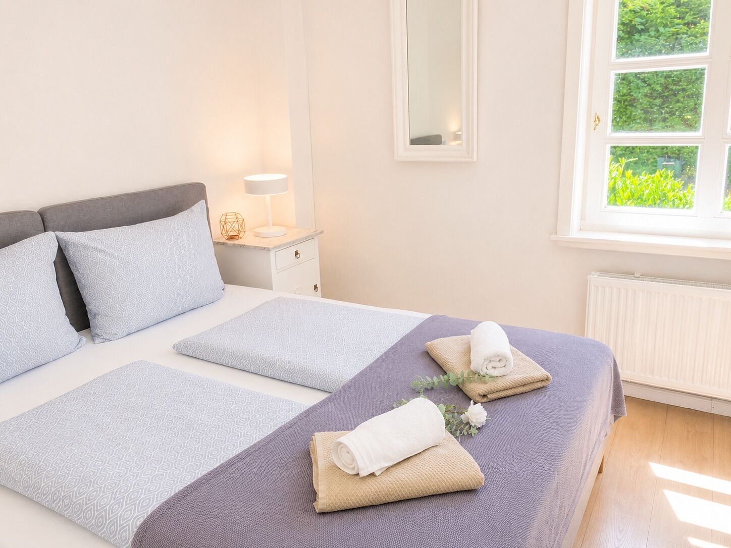 Ein ordentlich bezogenes Bett mit Kissen und einem gefalteten Handtuch, ein Nachttisch mit Lampe und ein Fenster mit Blick ins Grüne.