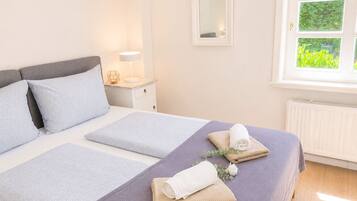 Ein ordentlich bezogenes Bett mit Kissen und einem gefalteten Handtuch, ein Nachttisch mit Lampe und ein Fenster mit Blick ins Grüne.