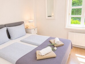 A neatly made bed with pillows and a folded towel, a bedside table with a lamp, and a window with a view of greenery.