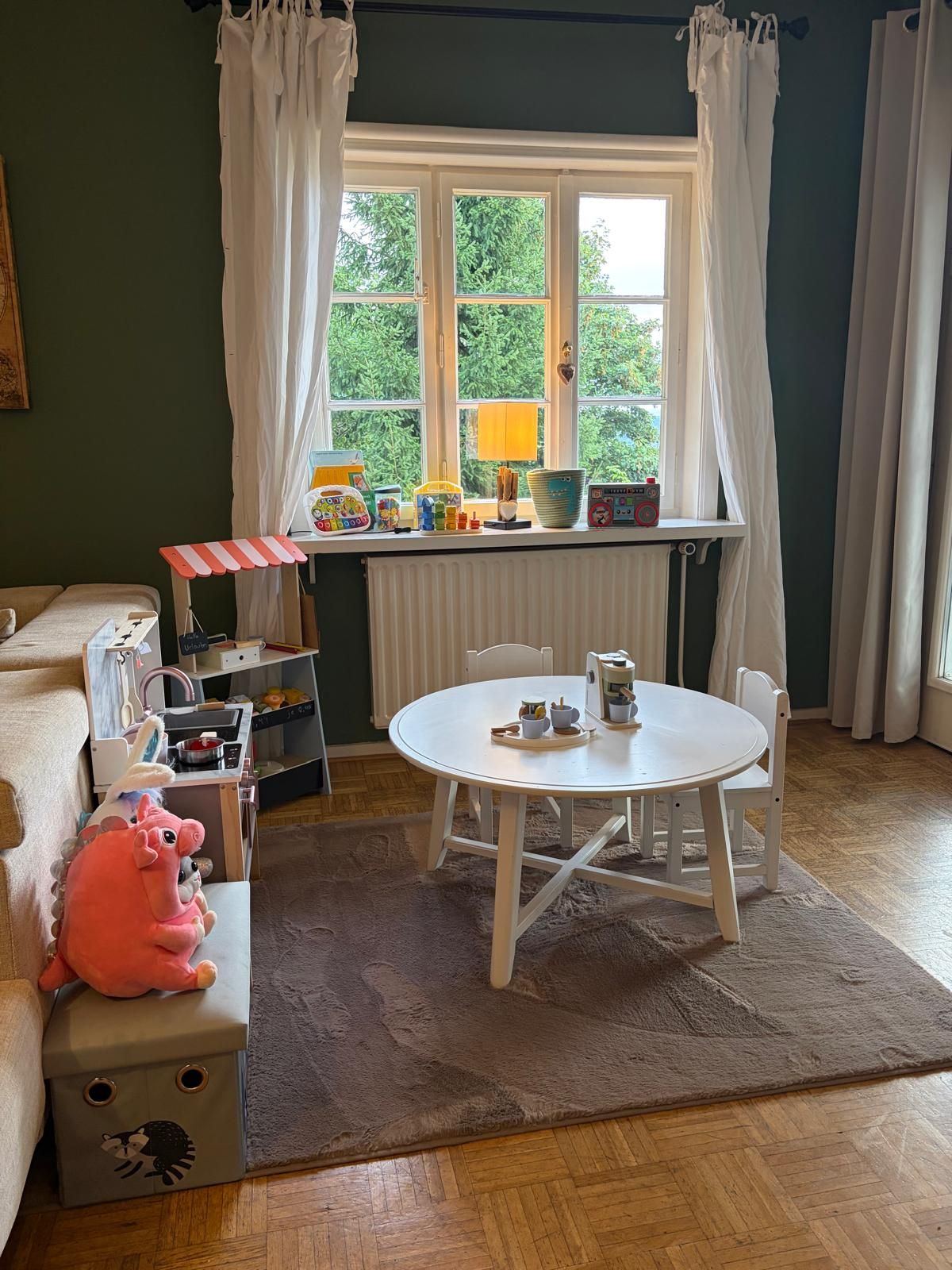 A cozy room with a round table, chairs, a radiator, a window with curtains, and a small play area with toys.