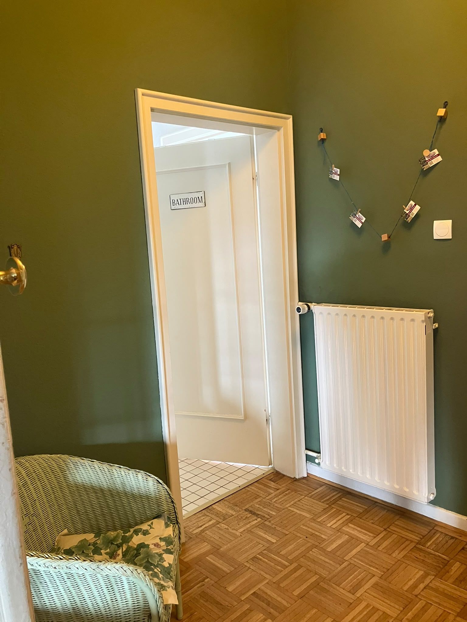 Ein Zimmer mit einer Tür, auf der 'BATHROOM' steht, einem Heizkörper, einem Fischgrätparkett und einem Korbstuhl.