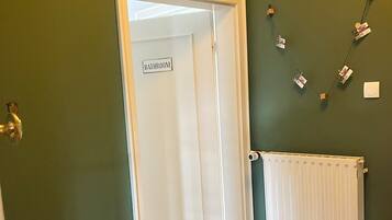 Ein Zimmer mit einer Tür, auf der 'BATHROOM' steht, einem Heizkörper, einem Fischgrätparkett und einem Korbstuhl.