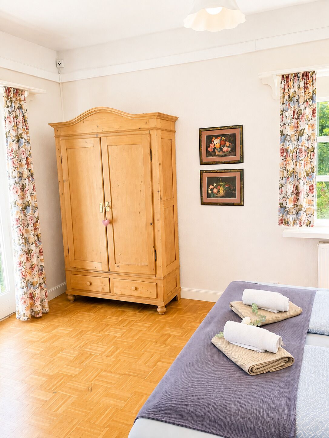 Una habitación con un armario de madera, dos cuadros enmarcados, una cama con toallas y cortinas estampadas.