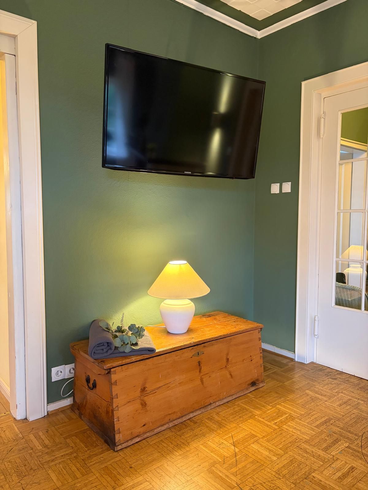 A room with a television mounted on the wall, a wooden chest of drawers, and a lamp on top.