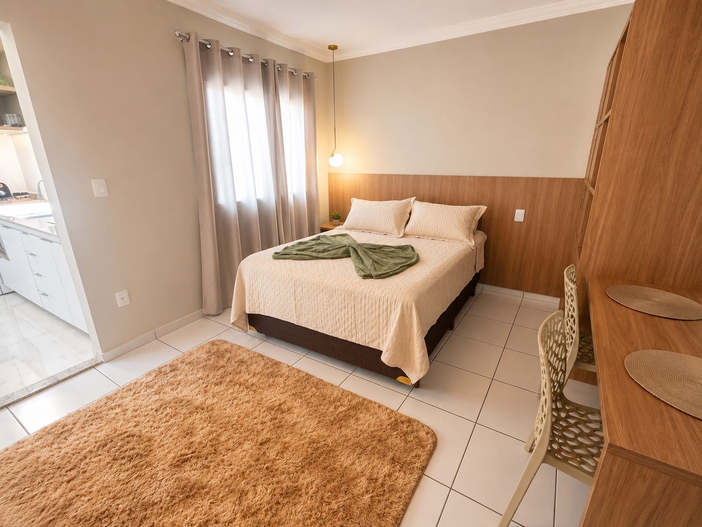 A bedroom with a bed, a nightstand, a lamp, a chair, and a view of the kitchen.
