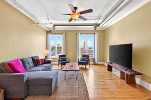 A living room with a ceiling fan, a flat-screen TV, a sofa, two chairs, and a coffee table. - Historic Downtown Loft | Steps from S&W Food Hall & Drum Circle! (Asheville)