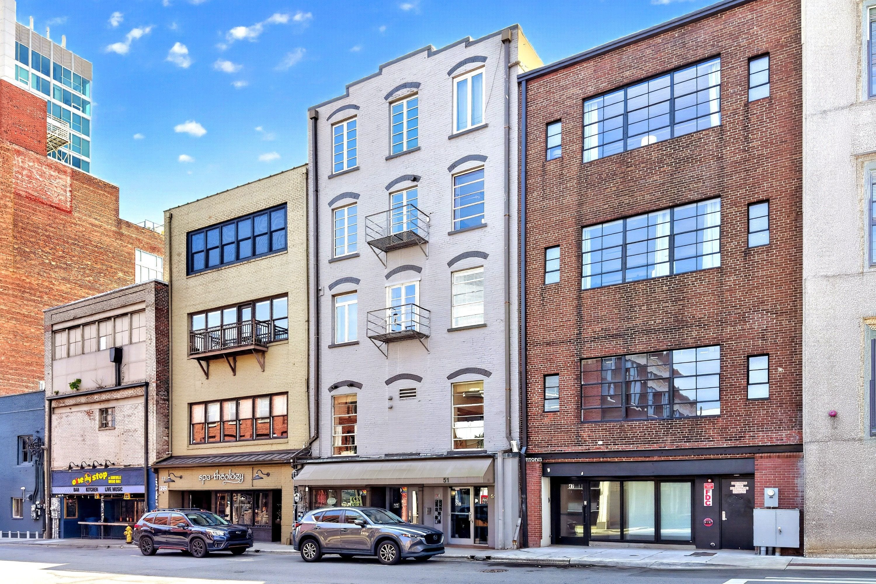 A multi-story building with a mix of brick and white facades, storefronts, and parked cars on the street.