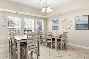 A dining area with a wooden table and chairs, large windows with blinds, and artwork on the walls. - Six by the Ocean - Spacious 6BR Condo w/ Shared Pool and Hot Tub (Sleeps 16) (North Myrtle Beach)