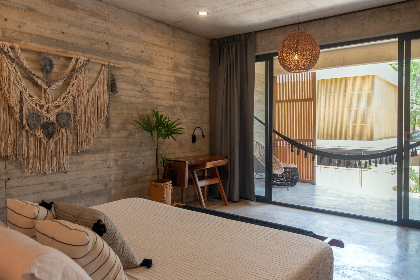 Una camera da letto con un letto, una scrivania, una sedia, un arazzo macramè e vista su una struttura in legno all'esterno.