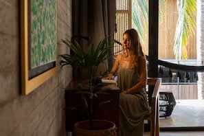 A person seated at a wooden table with a plant, in a room with a large window and a view of palm trees.
