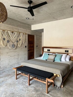 A modern bedroom with a bed, wooden bench, macramé wall decor, and a ceiling fan.