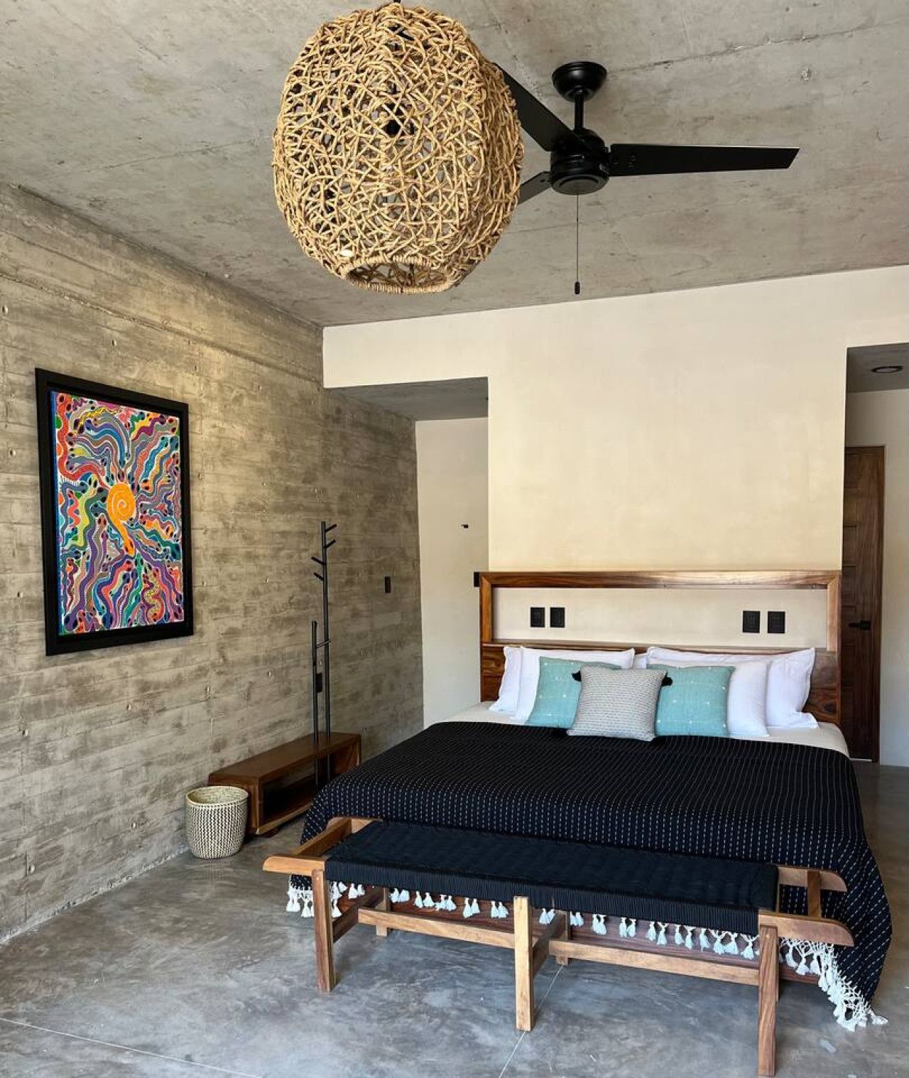 Een slaapkamer met een bed, een plafondventilator, een geweven lamp en een kleurrijk abstract schilderij aan de muur.