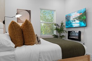 Ein Schlafzimmer mit einem Bett, einer Nachttischlampe, einem Fenster mit Jalousien, einem an der Wand montierten Fernseher und einem Bilderrahmen.