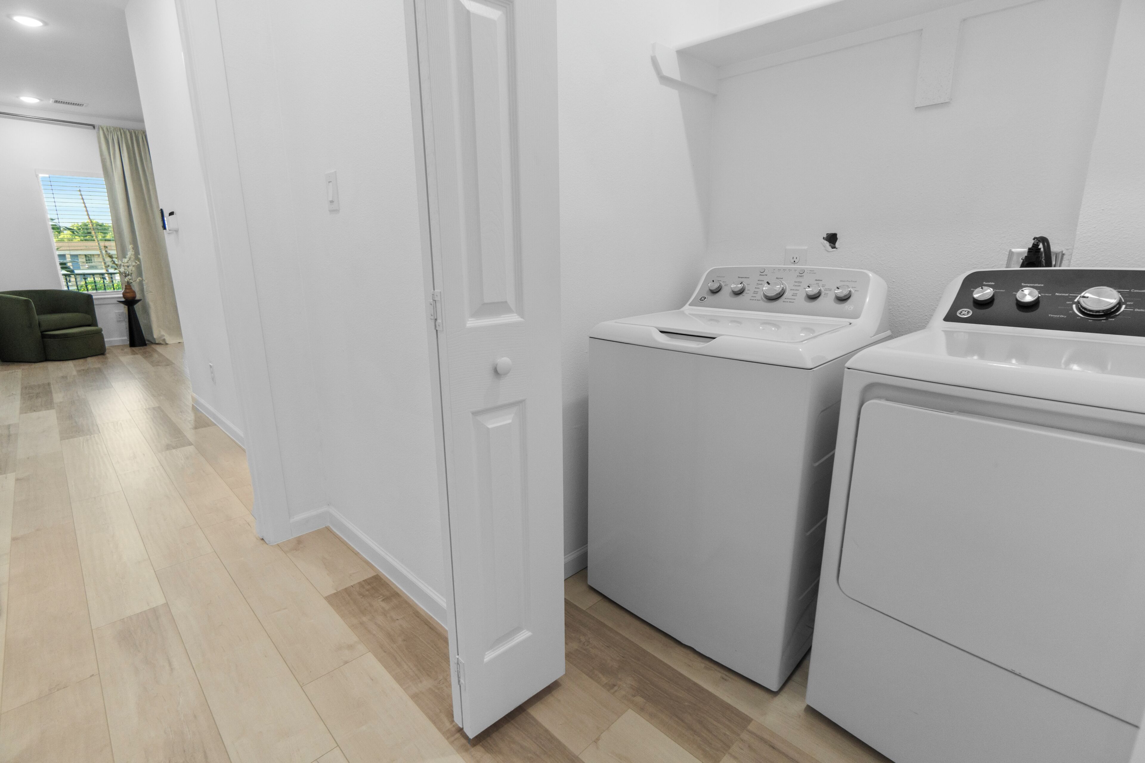 Un lavadero con lavadora y secadora, una puerta que da a otra habitación y vistas a un balcón con una planta.