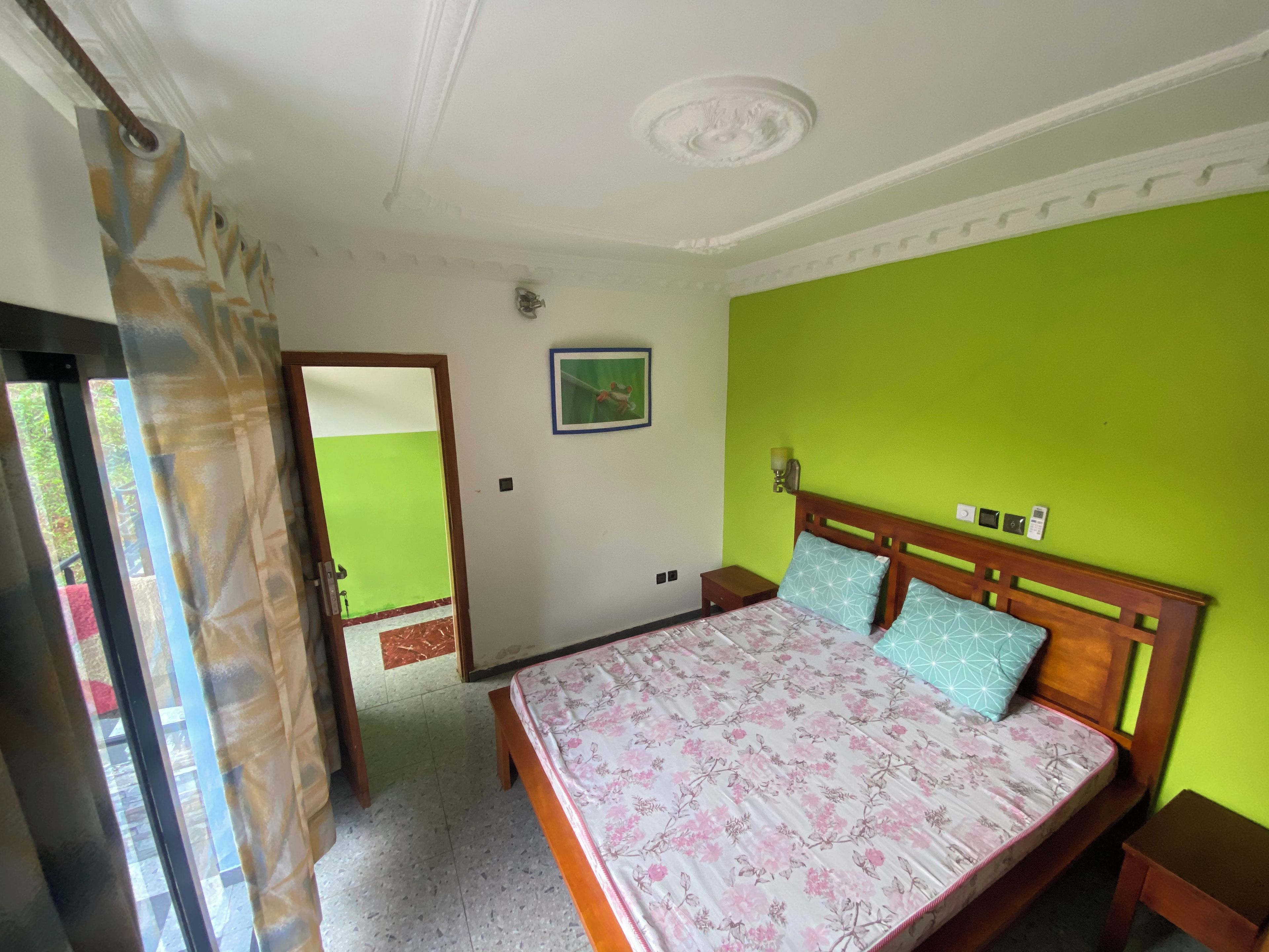 Quarto com cama, mesa de cabeceira, luminária na parede e porta para outro espaço.