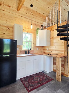 A kitchen with wooden walls, a black refrigerator, white cabinets, a red rug, and a hanging light fixture. - Peaceful 1-bedroom cabin for a cozy stay (Sandstone)