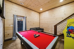 Ein Billardtisch mit roter Spielvliesbahn, ein graues Sofa und ein Fenster mit Vorhängen.