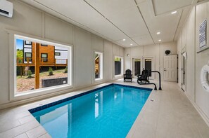A modern indoor swimming pool with clear windows, a wooden deck outside, and a poolside chair. - Brand new, indoor heated pool, hot-tub, closer to attractions, games, theater (Sevierville)