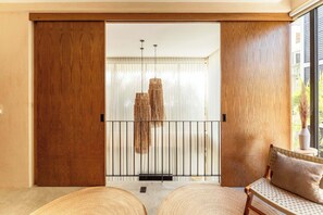 Ein Zimmer mit einer Schiebetür aus Holz, einem gewobenen Rattan-Sessel, einer gewobenen Fußmatte und einer Hängeleuchte. Blick ins Grüne außerhalb.