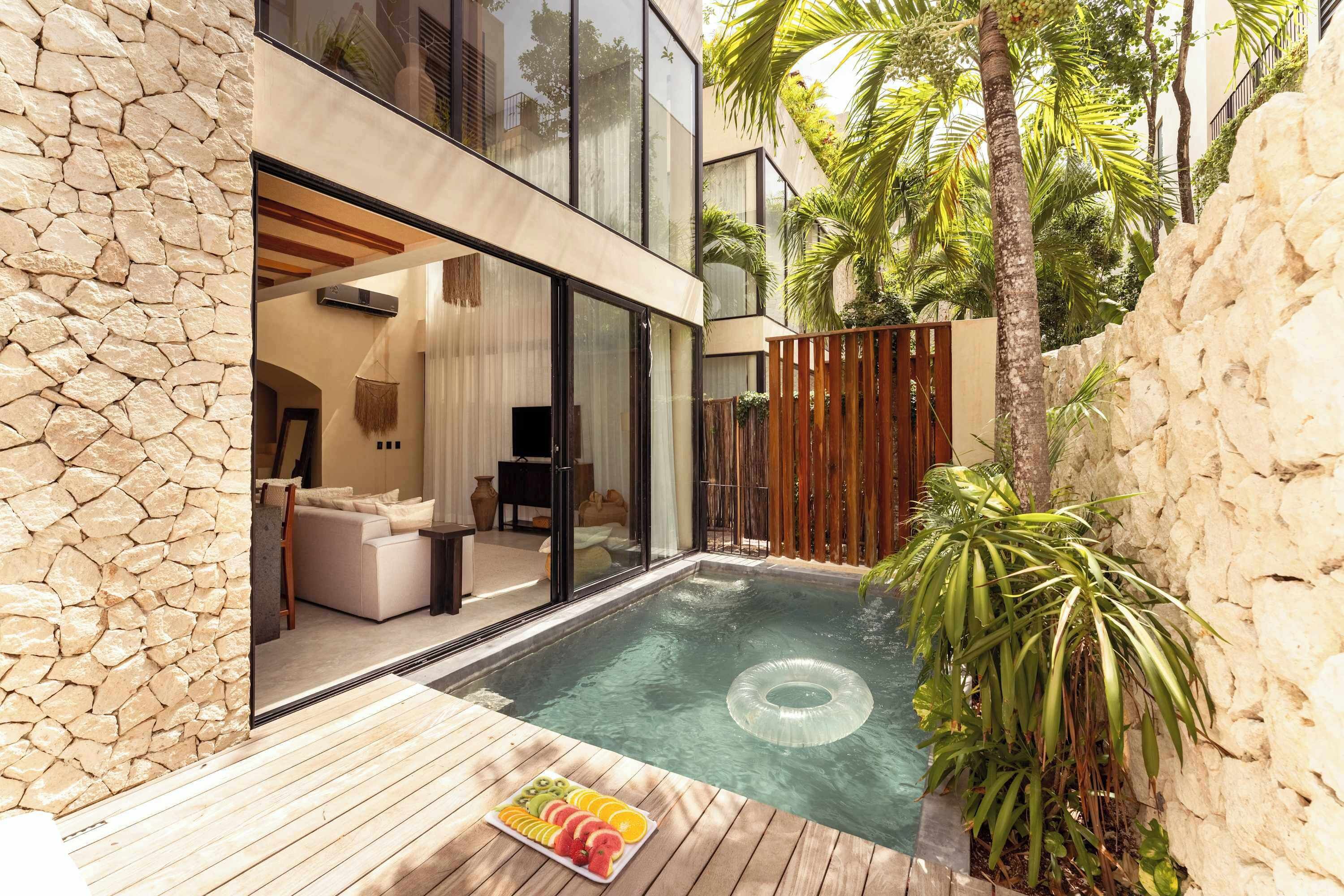 A modern outdoor pool area with a wooden deck, a stone wall, and a glass-enclosed living space with a sofa and a television.