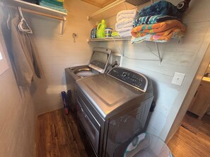 A laundry room with a front-loading washing machine, shelves with folded towels, and cleaning supplies. - Quaint 1-bedroom studio with AC in Oak Island - just a 1/2 mile to the beach! (Oak Island)