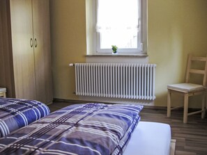 A bedroom with two beds, a radiator, a window with a plant, and a wooden chair. - Ferienwohnung Debacher by Interhome (Rust)