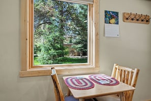 A room with a wooden table and chairs, a calendar on the wall, and a window with a view of trees and a brick wall. - 7 minutes to Yellowstone The Aspen Cabin (West Yellowstone)