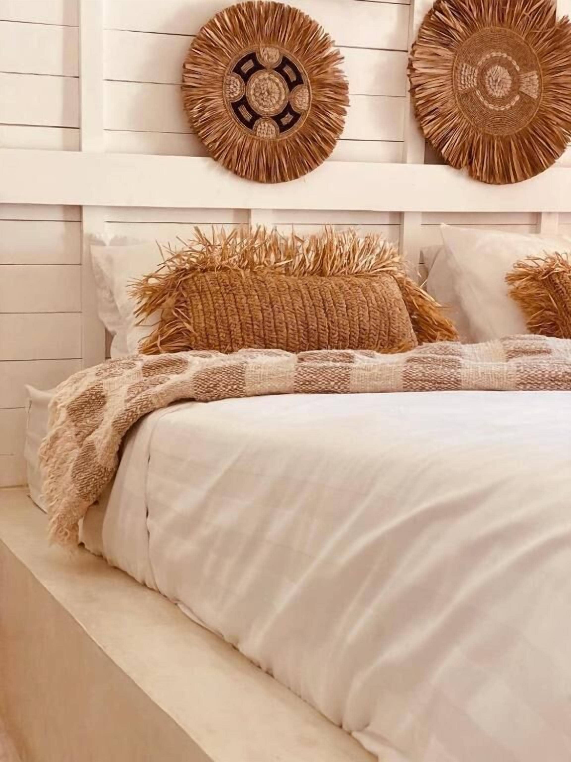 Ein Bett mit weißer Decke und zwei braunen Kissen, darüber ein gewebter Wandbehang und ein weißer Kopfteil.