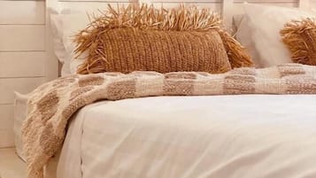 Una cama con edredón blanco y dos almohadas marrones, un tapiz tejido en la pared superior y un cabecero blanco.