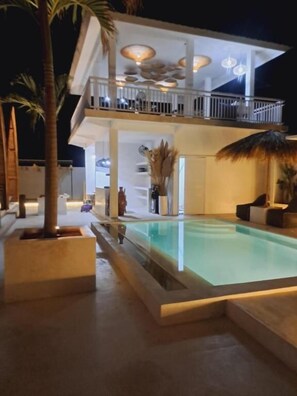 Un área de piscina con superficie de agua cristalina, una sombrilla de paja y un edificio de dos pisos con balcones.