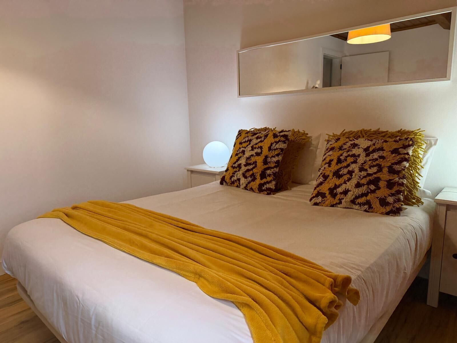 Een bed met een geel plaid, twee gestreepte kussens, een bedlampje en een ronde spiegel op een witte nachtkastje.