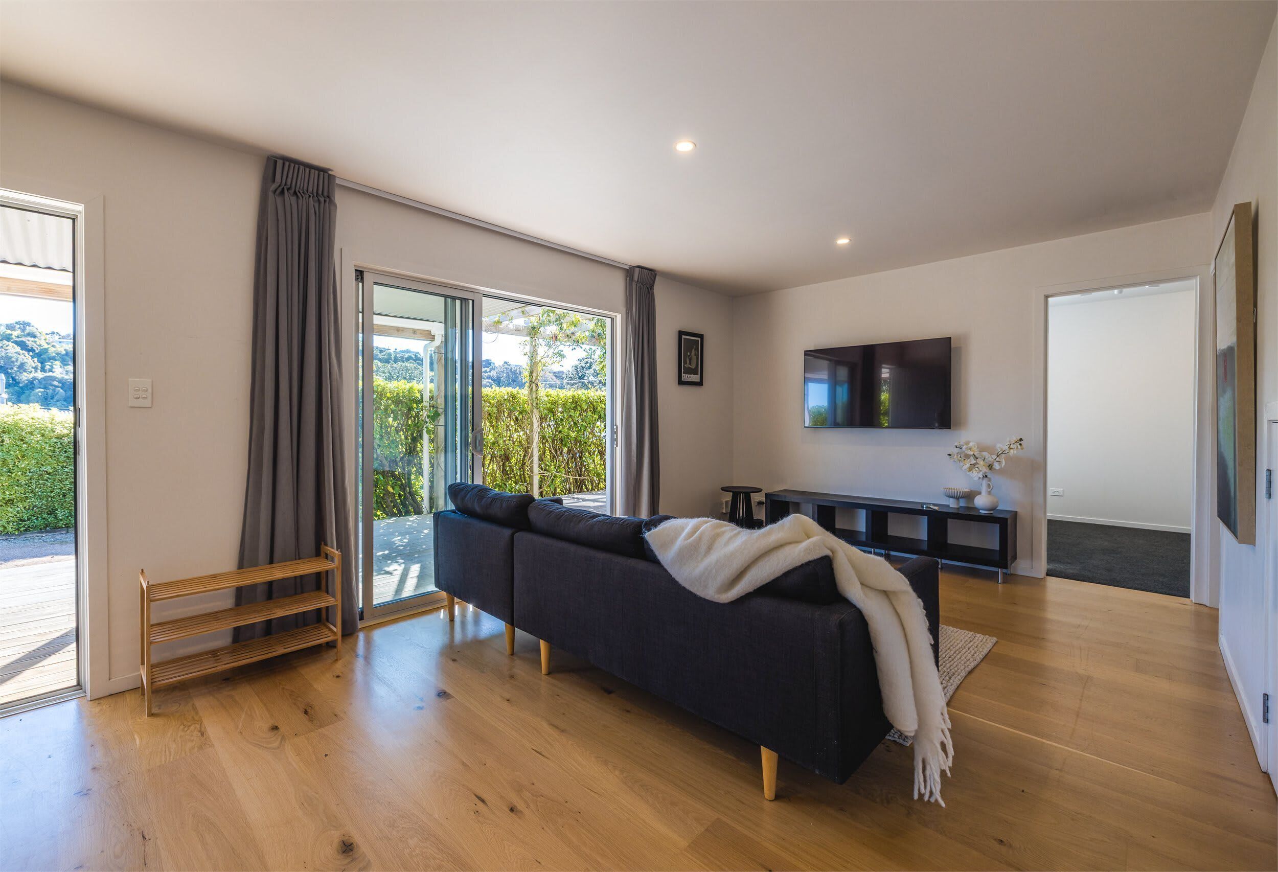 Ein modernes Wohnzimmer mit einem dunklen Sofa, Holzboden, einem an der Wand montierten Fernseher und einer Glastür zum Balkon.